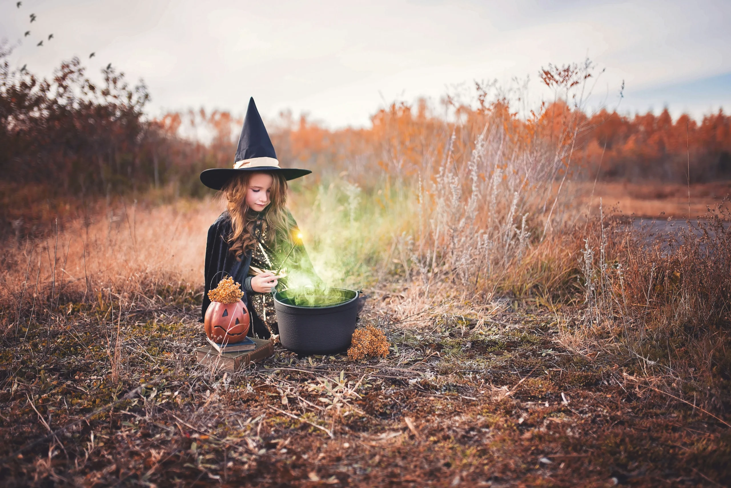 A child wearing a witch costume stirs a cauldron in a fall field with autumn colors