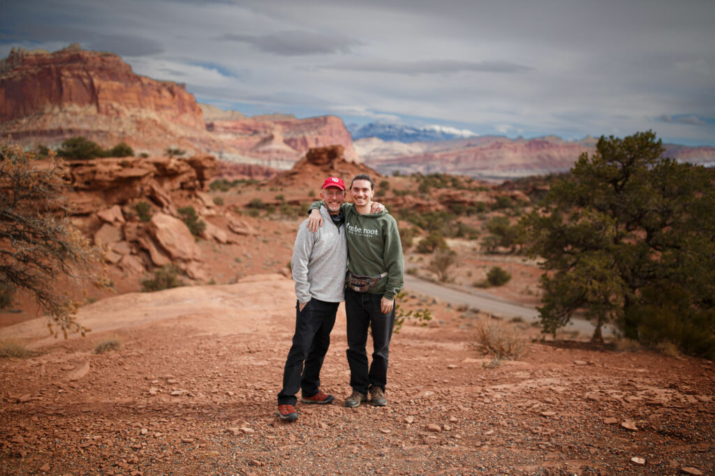 Two people stand arm in arm on a rocky desert overlook in Escalante Utah, smiling in front of sweeping red rock cliffs and a cloudy canyon landscape.