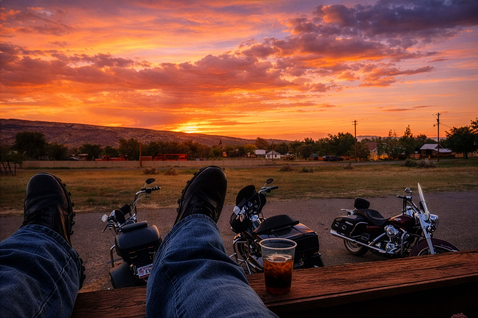 View from Escalante Canyon Inn at sunset, with a guest’s boots up on the porch railing, an iced drink, and three parked motorcycles against a vivid desert sky.